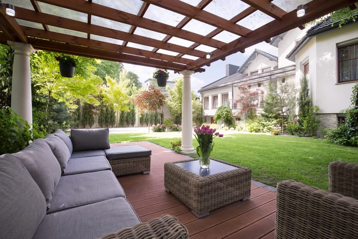 Terrazza coperta con pergolato in legno, arredi da esterno in vimini, tavolo con vaso di fiori freschi e splendida vista sul giardino curato con casa sullo sfondo.