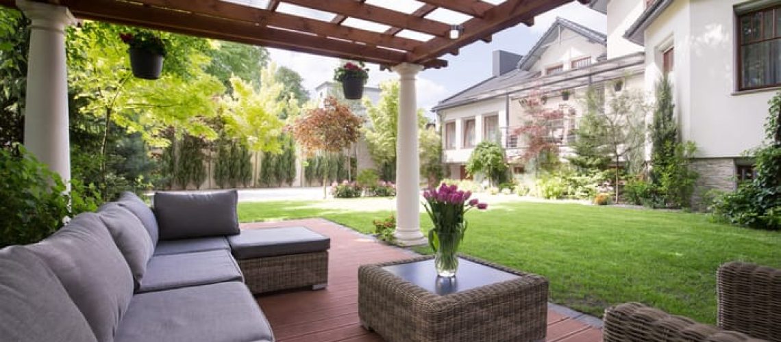 Terrazza coperta con pergolato in legno, arredi da esterno in vimini, tavolo con vaso di fiori freschi e splendida vista sul giardino curato con casa sullo sfondo.